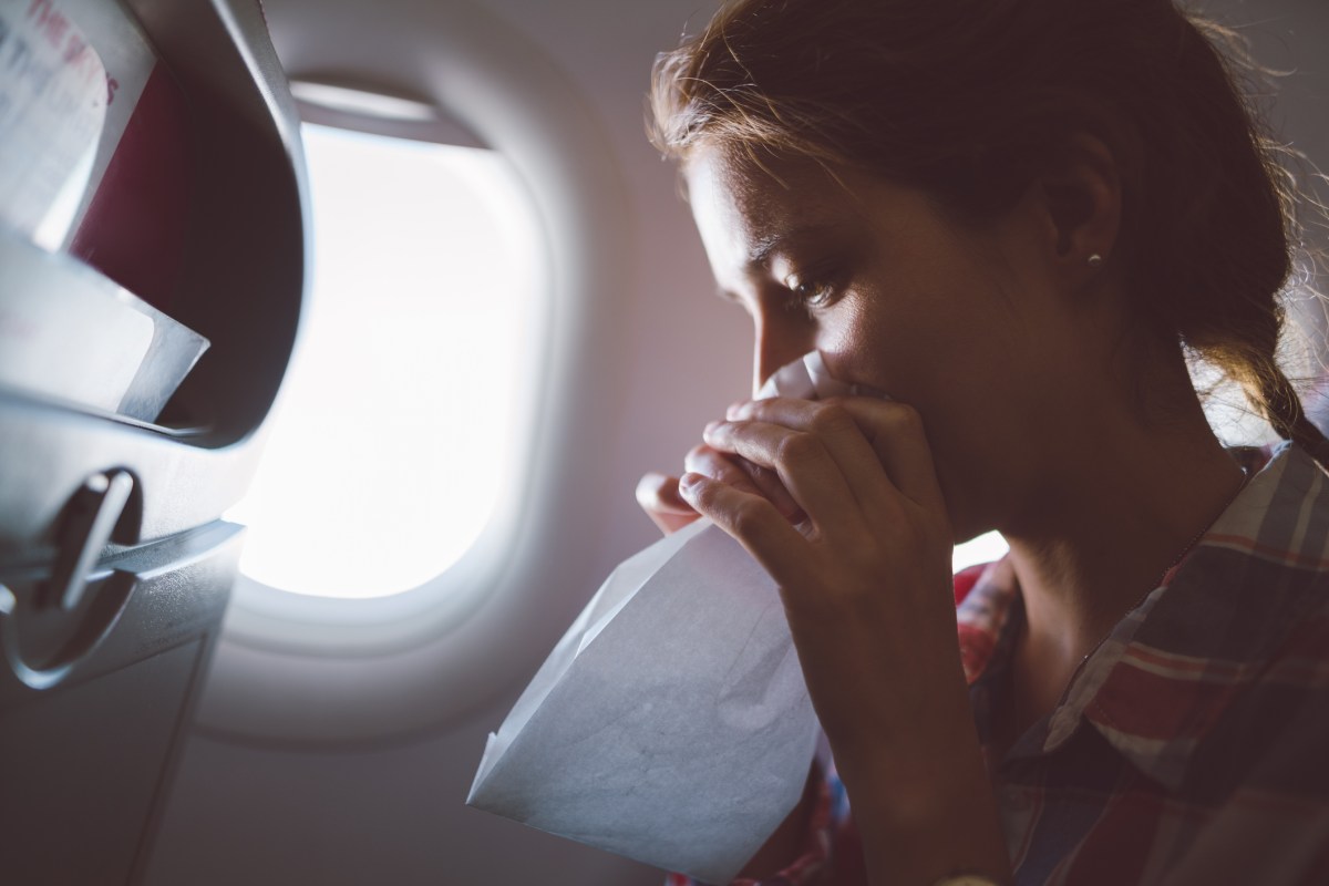 Frau mit Reiseübelkeit sitzt im Flugzeug, sie hält eine Papiertüte vor dem Gesicht