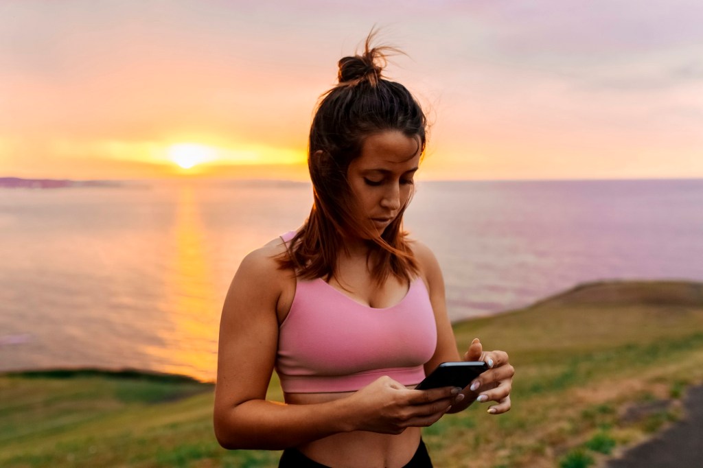 Frau vor dem Sonnenuntergang im Sport-BH mit Smartphone in der Hand