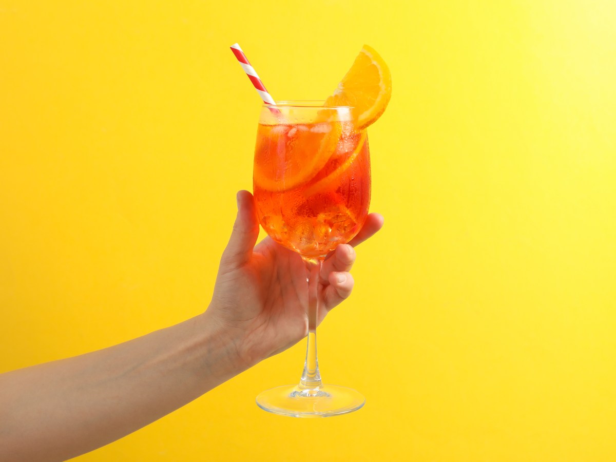 Aperol Spritz im Glas mit Strohhalm in der Hand auf gelbem Hintergrund