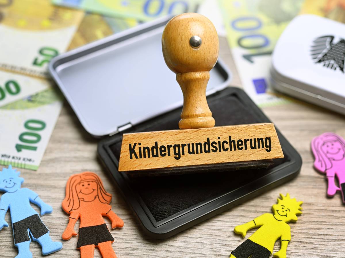 Stempel mit der Aufschrift "Kindergrundsicherung" liegt zwischen Geldscheinen und Kinderfiguren.