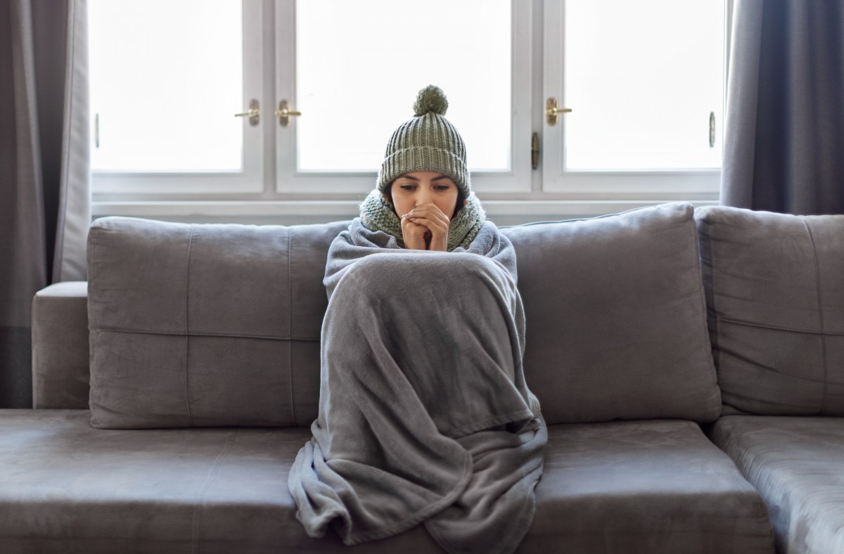 Eine Frau sitzt mit einer decke auf dem Sofa. Sie trägt eine Münze und friert.