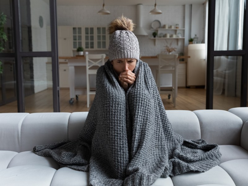 Eine Frau sitzt frierend in ihrer Wohnung. Sie trägt eine Mütze und ich in eine Decke eingewickelt.