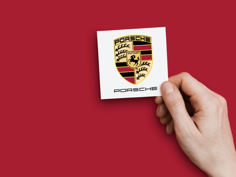 Eine Hand hält das Logo von Porsche vor eine rote Wand.