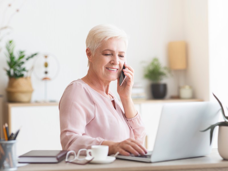 Eine ältere Dame sitzt am Laptop und telefoniert.