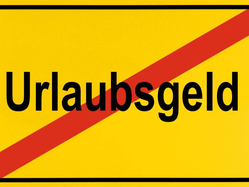 Auf einem gelben Ortsschild steht das Wort "Urlaubsgeld". Es ist durchgestrichen.