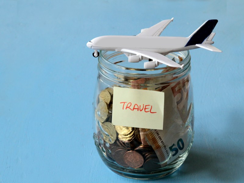 In einem Glas sind viele Münzen. Auf ihm liegt ein Flugzeug und ein Zettel mit der Aufschrift "Travel" klebt am Glas.