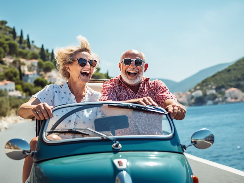 Ein Seniorenpaar sitzt im Cabrio und fährt an der Küste entlang.