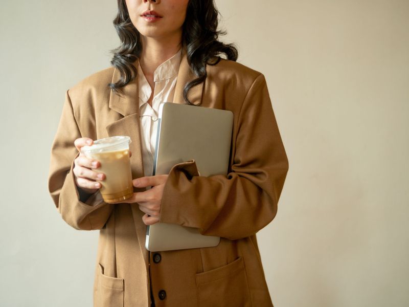 Eine Frau im beigen Mantel hält ihren Laptop und einen Eiskaffee in der Hand.