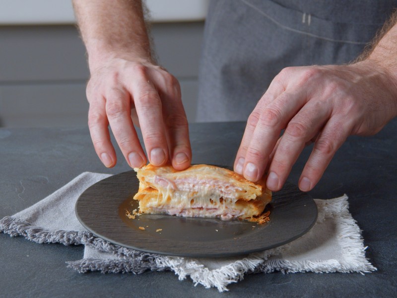 Käse-Schinken-Toasties aus Blätterteig auf einem Teller, männliche Hände drücken sie zusammen.