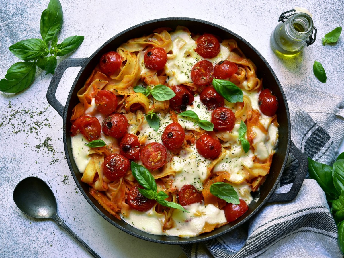 Nudelauflauf mit Tomaten und Mozzarella in runder Auflaufform, garniert mit Basilikum, daneben ein Löffel. Draufsicht.