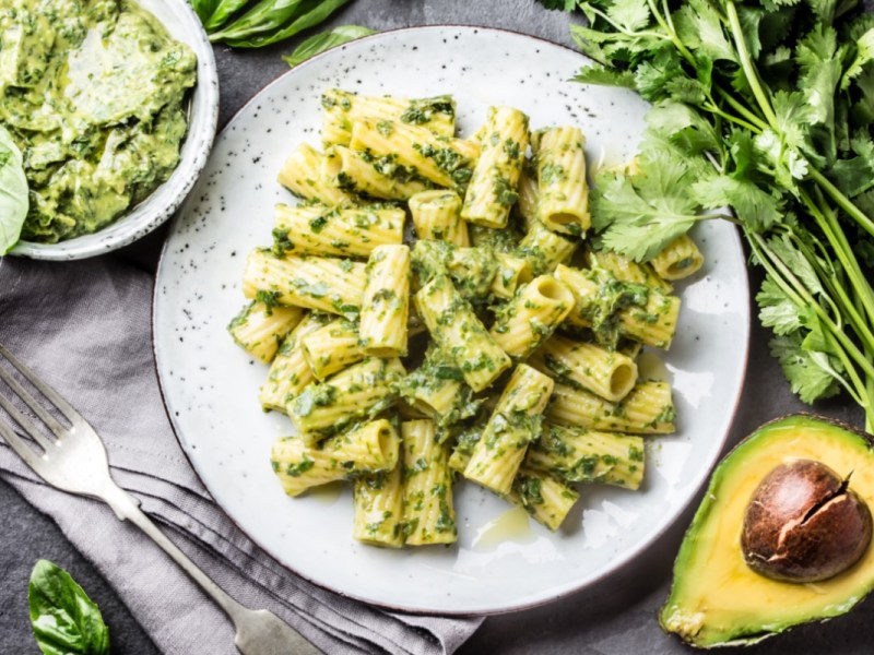 Ein Teller Nudeln mit Avocado-Pesto in der Draufsicht. Daneben eine Schüssel Pesto und frische Zutaten.