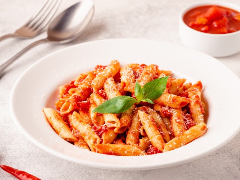 Ein Teller Penne all’arrabbiata. Daneben steht ein Schälchen mit Tomaten und es liegt Besteck links vom Teller.