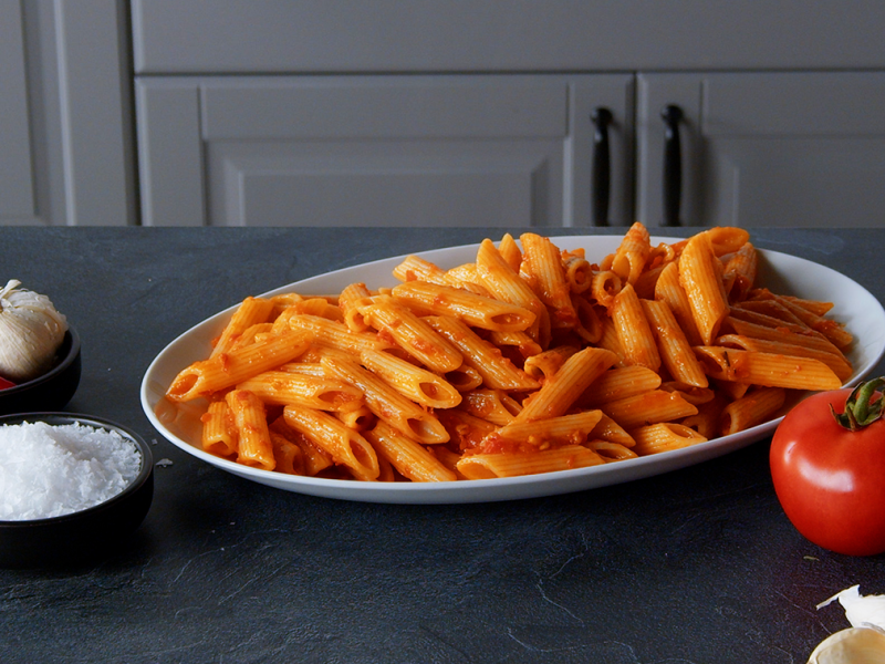 Penne Arrabiata, angerichtet auf einem Teller. Tomaten und Gewürze liegen außen herum.