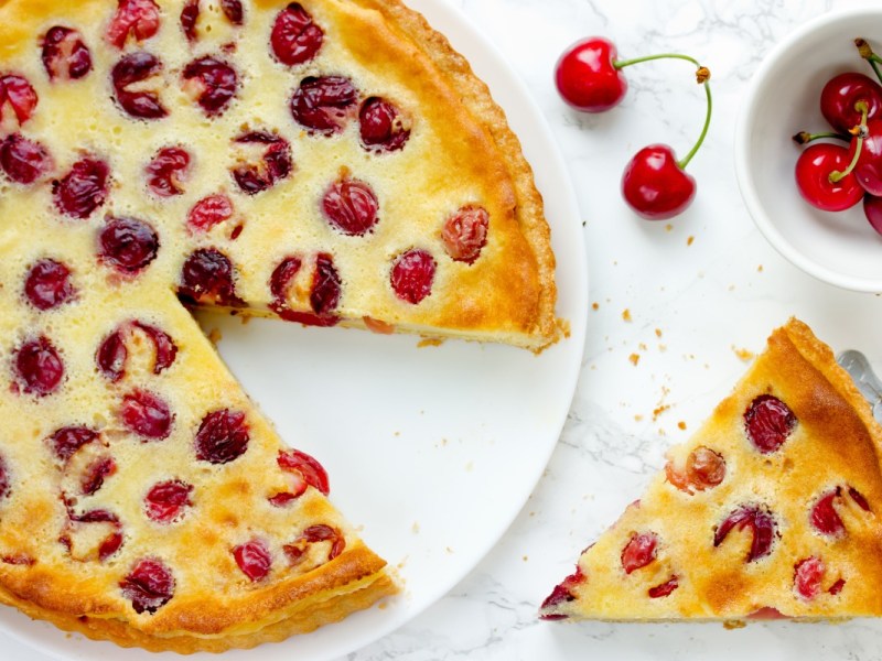 Ein Pudding-Kirschkuchen, aus dem ein Stück geschnitten wurde, in der Draufsicht. Das Kuchenstück sowie eine Schale mit frischen Kirschen sind daneben.