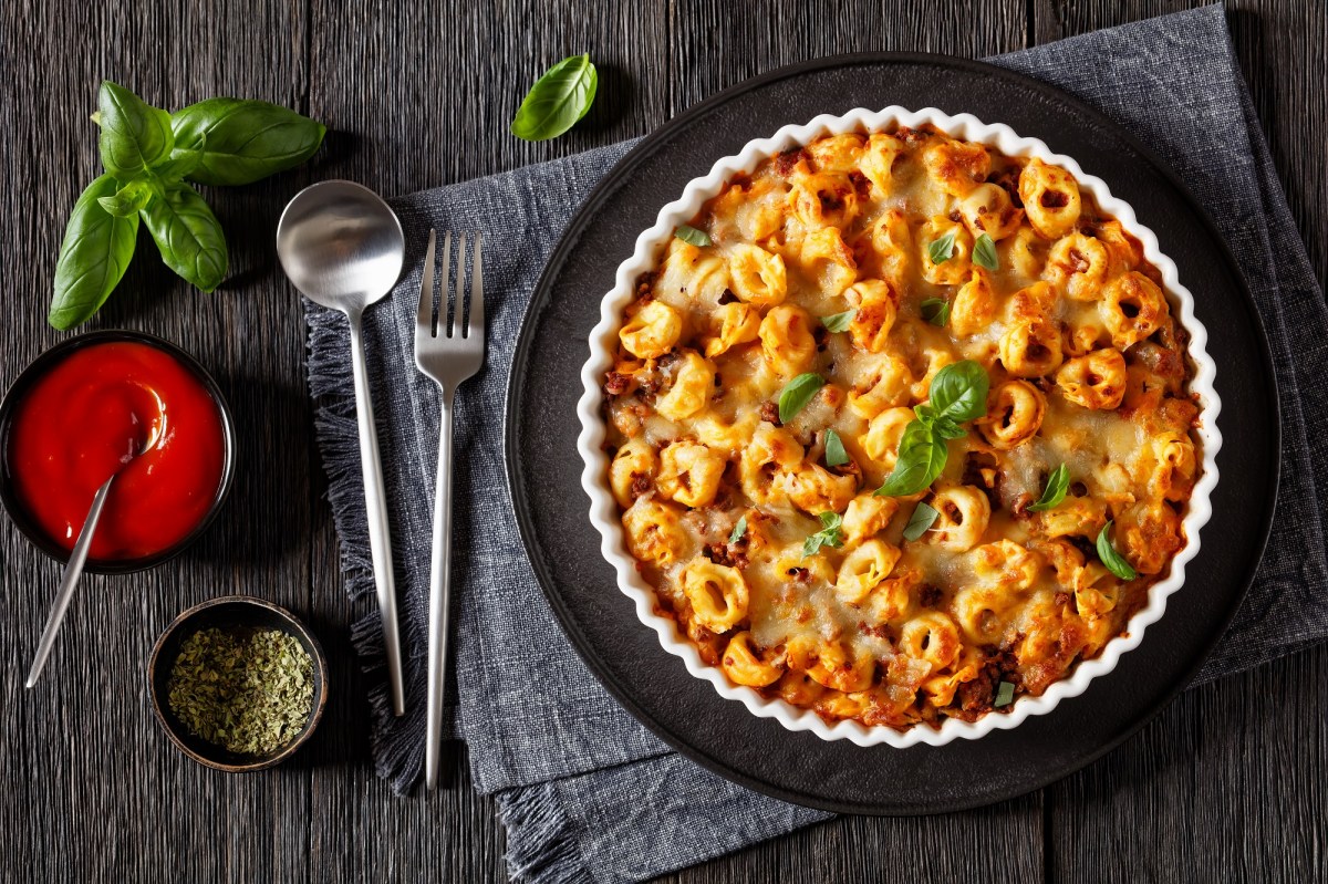 Tortellini-Auflauf in einer geriffelten Auflaufform auf einem schwarzen Tablett. Darunter ein graues Platzdeckchen, Löffel und Gabel und Tomatensoße. Draufsicht.