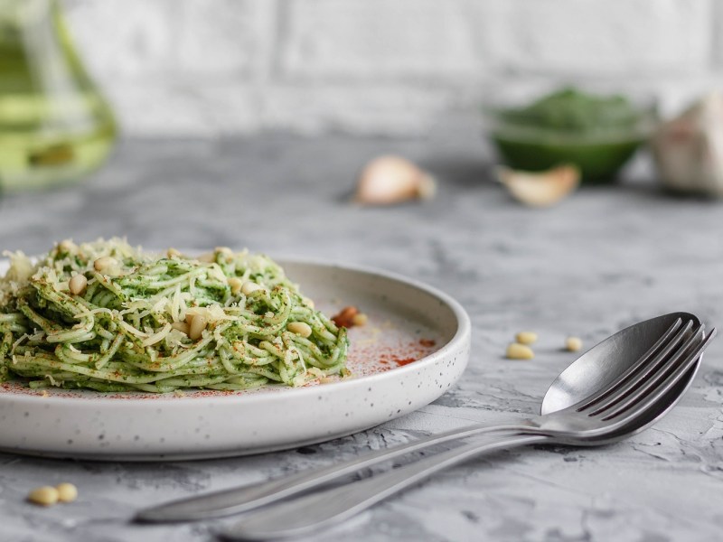 Ein Teller Zucchini-Avocado-Pasta garniert mit Pinienkernen und Parmesan, daneben ein Löffel.