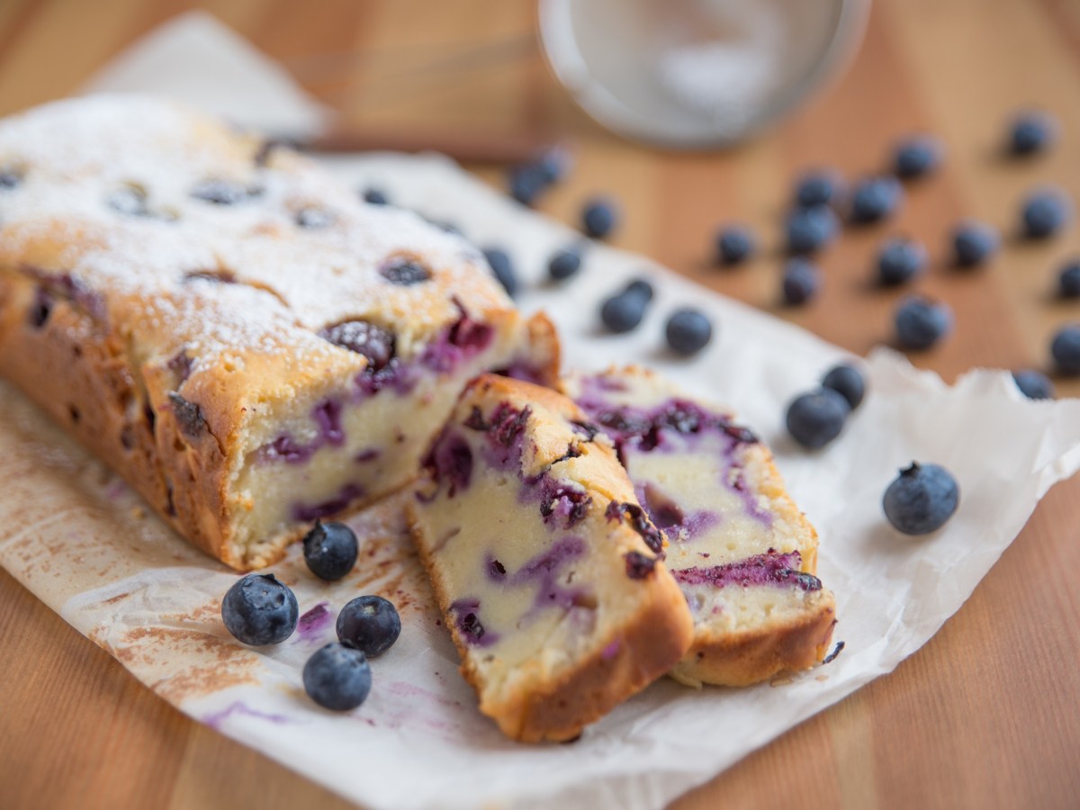 Blaubeer-Joghurt-Kuchen auf Backpapier mit zwei abgeschnittenen Stückchen.