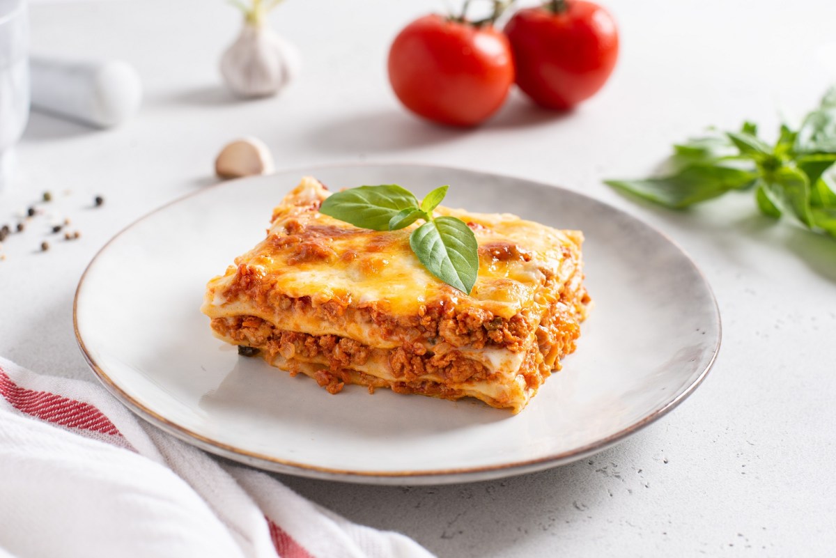 Ein Stück Lasagne mit Schmand auf einem Teller, garniert mit Basilikum, dahinter 2 Tomaten.