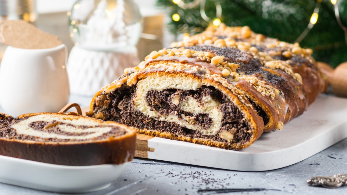 Ein aufgeschnittener Mohnstriezel mit Walnüssen und Streuseln, der auf einer länglichen Servierplatte liegt. Links davor liegt ein Stück auf einem kleinen Teller. Im Hintergrund sind ein Milchkännchen, eine Schneekugel und Tannengrün mit einer Lichterkette zu sehen.