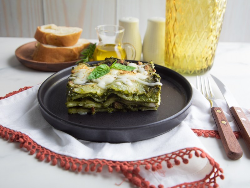 Pesto-Lasagne auf einem schwarzen Teller. Dahinter stehen ein Teller Brot, eine Flasche Olivenöl und Salz nud Pfeffer.