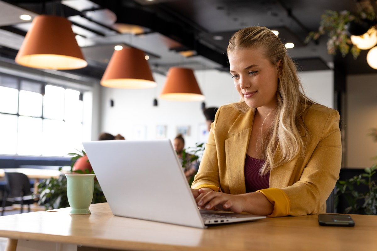 Eine Frau mit blonden Haaren sitzt im Büro und arbeitet am Laptop.