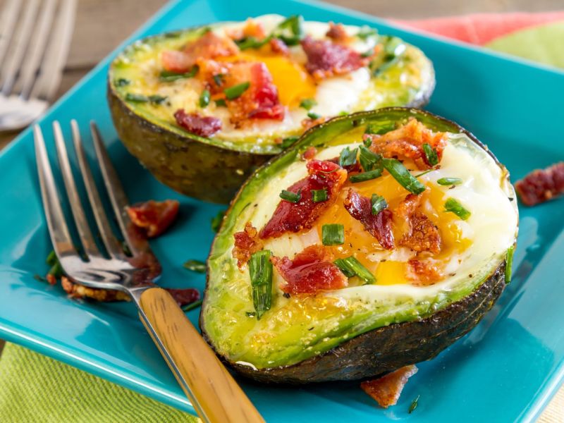 Aufgeschnittene Avocado-Eier, gefüllt mit Bacon und Kräutern. Eine gelbe Gabel liebt neben dem Teller.