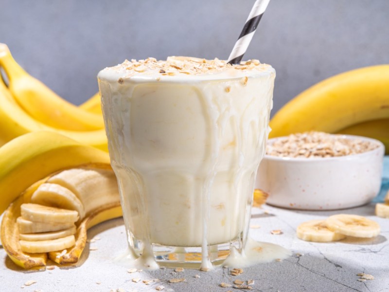 Ein Glas Frühstücksshake mit Bananen und Haferflocken. Im Hintergrund sind Bananen uds eine Schale mit Haferflocke zu sehen.