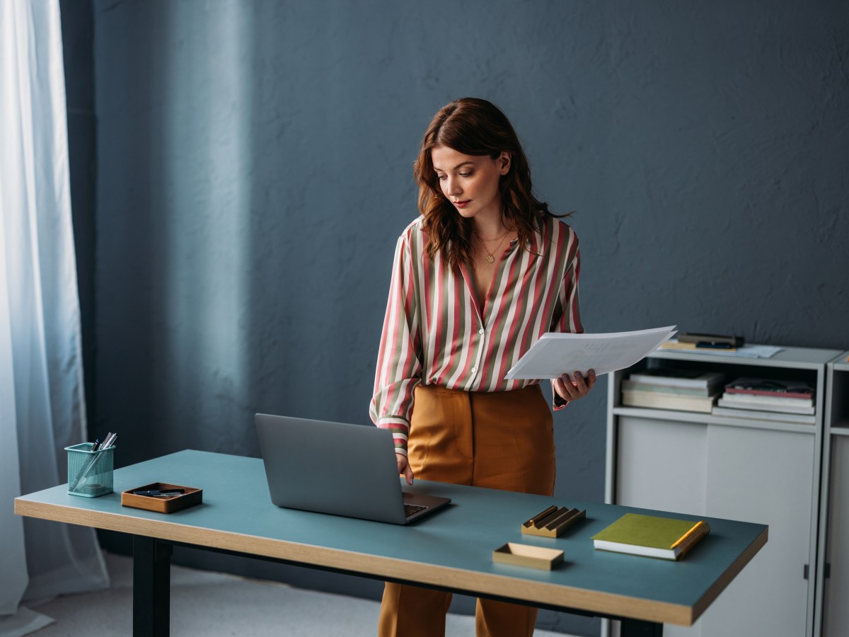 Konzentrierte, berufstätige Frau steht mit Laptop und Papierkram an einem Schreibtisch in einem modernen Büroarbeitsplatz.
