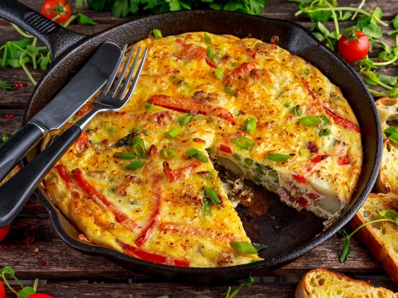 Quiche-Auflauf, von dem ein Stück fehlt, in einer gusseisernen Pfanne. Darauf liegen Messer und Gabel und drumherum ist der Tisch mit Brot und Rucola bedeckt.