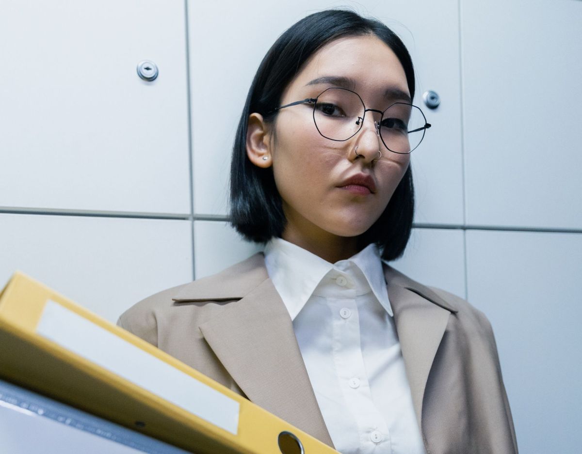 Eine Frau mit einem schwarzen Bob und Brille trägt einen gelben Aktenordner.