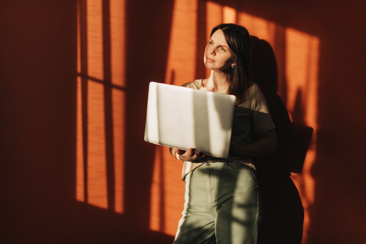 Eine Frau mit einem Laptop auf dem Hintergrund einer Terrakottawand, die Sonne scheint hell.