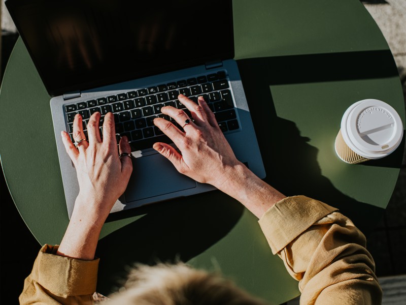 Eine Frau arbeitet draußen an ihrem Laptop. Neben dem PC steht ein Kaffee to go.