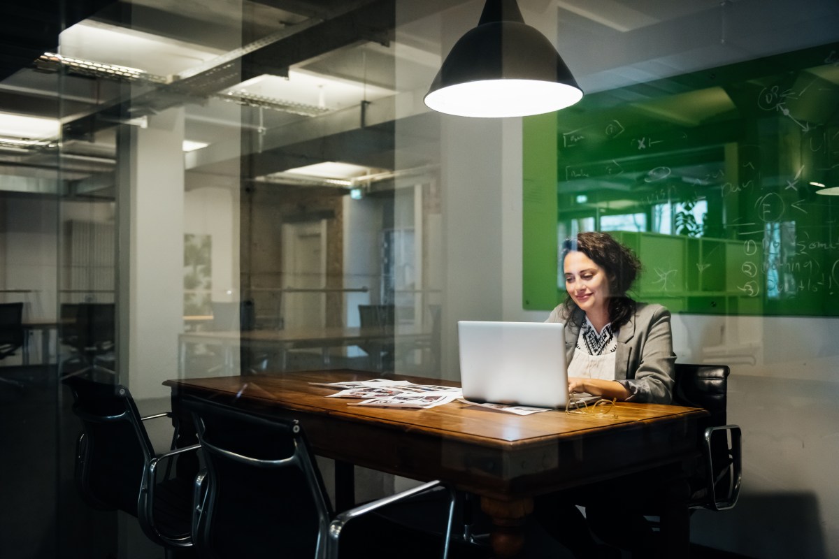 Eine Frau sitzt an einem Tisch in einem Büro und blickt zufrieden auf einen Laptop.
