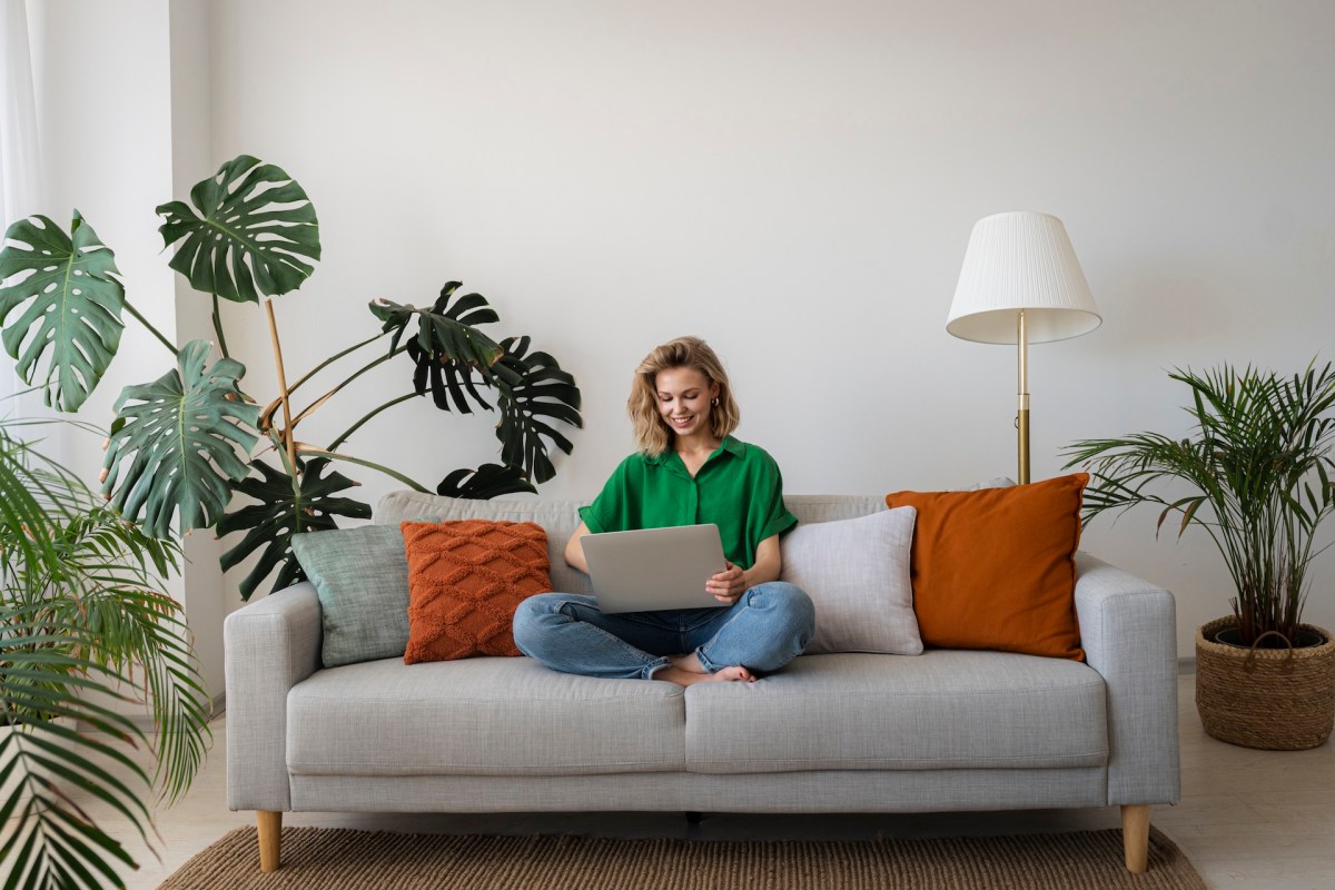 Eine junge Frau sitzt zu Hause auf dem Sofa und arbeitet am Laptop.